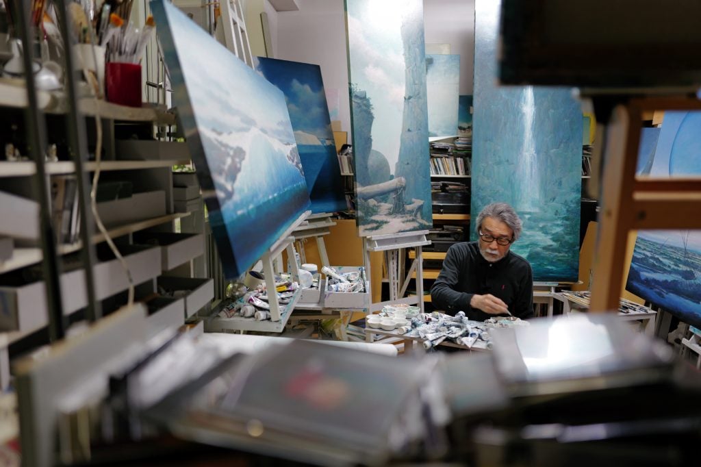 This image shows a bespectacled East Asian man in grey hair and beard painting in an artist studio, surrounded by multiple paintings in blue.