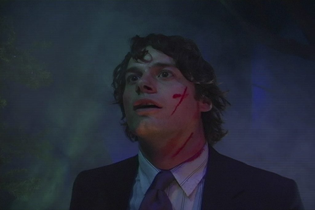 a young white man in a suit looks to the corner of the ceiling with a splotch of red blood on his cheek