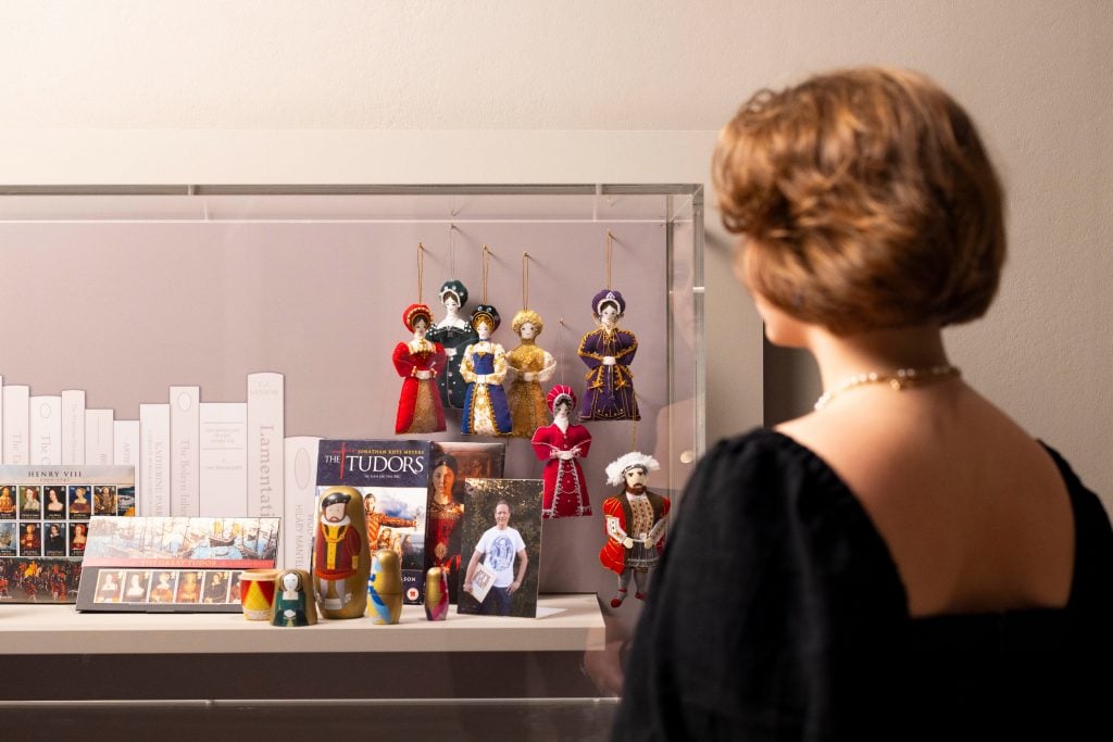 a woman standing in front of a display case filled with little figurines, ornaments, books etc that are in some related to the Six Wives of Henry VIII