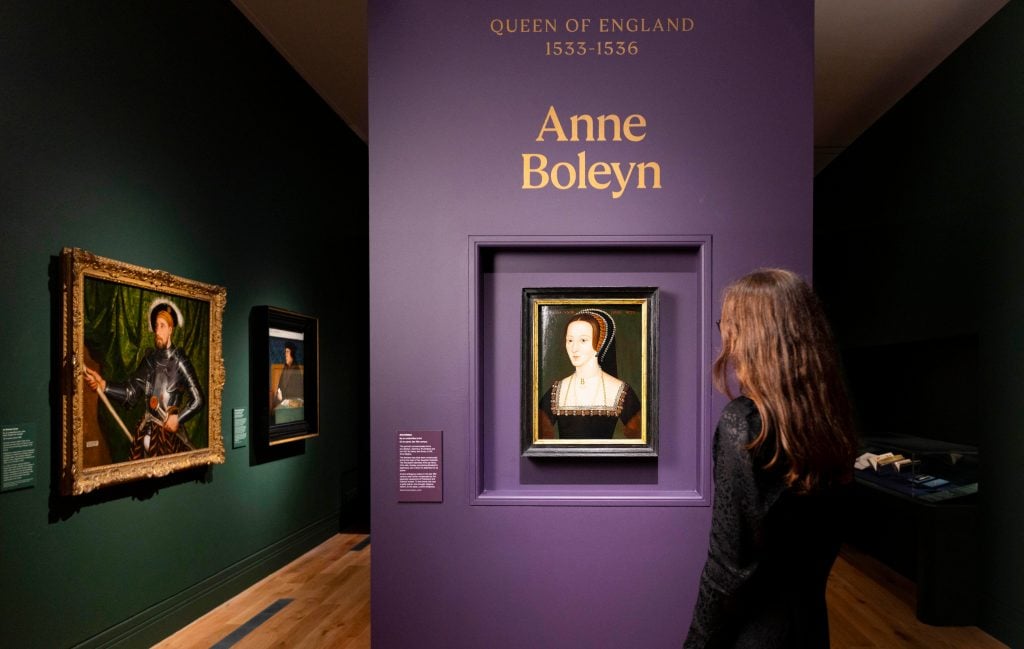 an interior of a museum show in which a purple wall is hung with a portrait of a woman in the TUdor style and above in gold letters is written 
