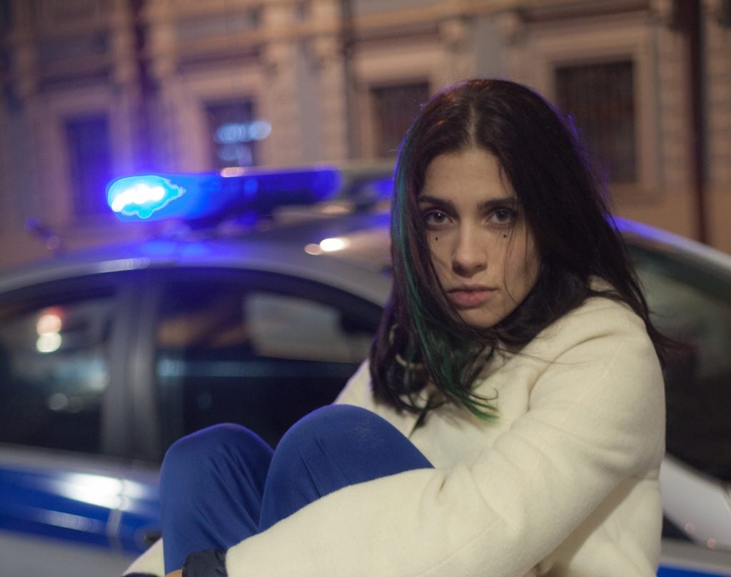 a woman in a white coat hugs her knees and stares intensely at the camera. Behind her you can see the blue lights of a police car