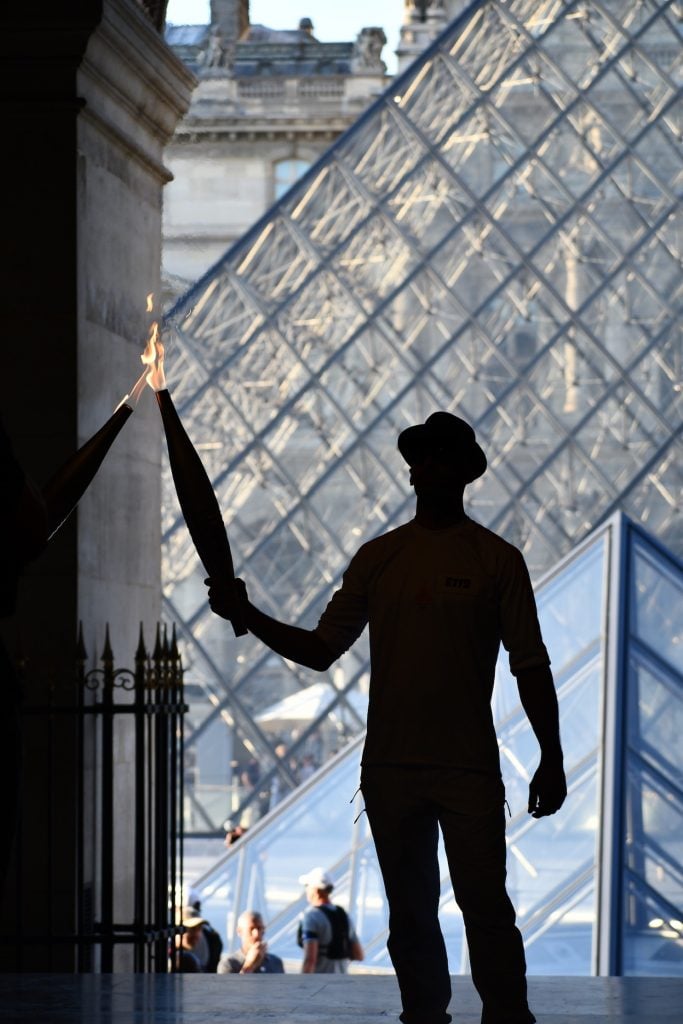The silhouette of a man in a fedora holding a lit torch passes the flame to another torch seen coming out of a doorway. Behind him a is a stone wall with molding and a partial view of two glass paneled pyramids.