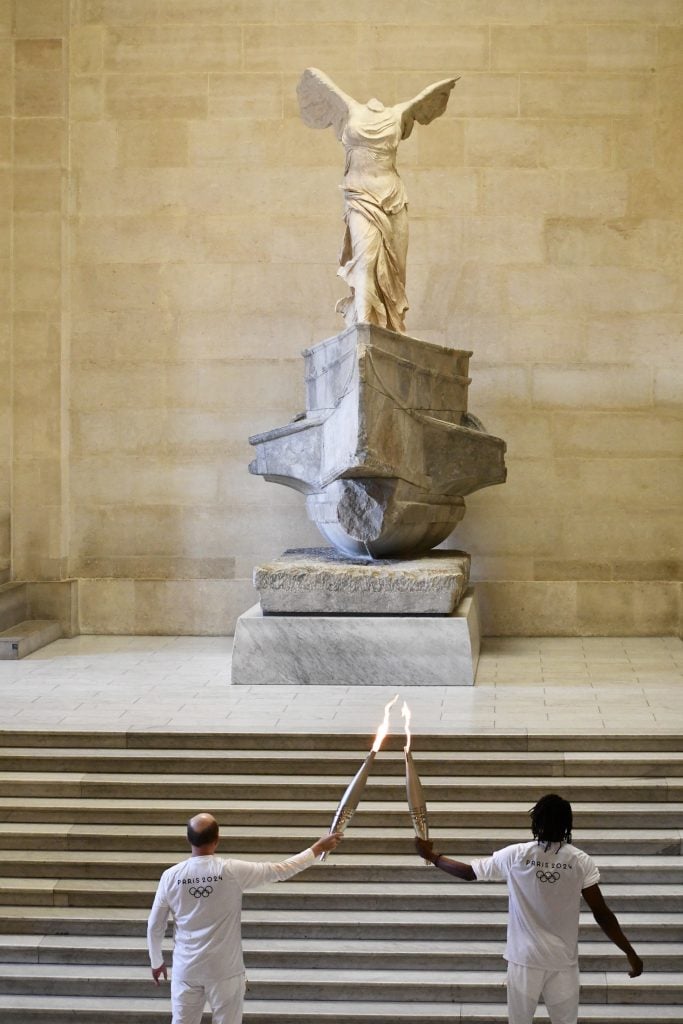 Two men seen from behind pass the flame of the Olympic torch on the stairs in front of an armless classical marble statue of a winged women in a draped robe that stands on a large pedestal in front of a white marble wall in a museum.