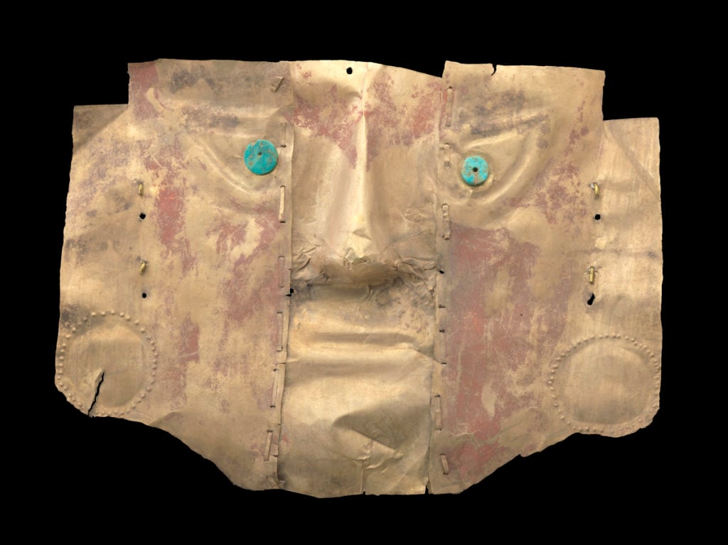 An ancient mask made of metal with a human-like face, featuring turquoise eyes and a weathered, reddish patina, indicating age and historical significance.