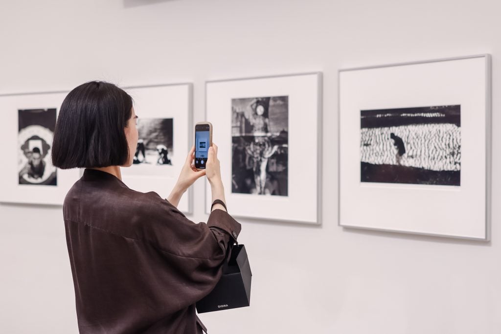 A woman in dark hair dressed in a blouse in dark colour is taking photos of photography works on the wall with a smart phone.