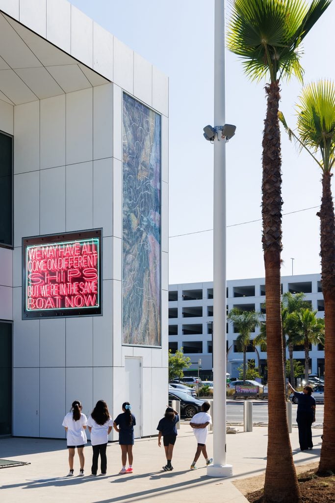 A group of children walk past a modern building featuring a neon sign that reads, “WE MAY HAVE ALL COME ON DIFFERENT SHIPS BUT WE’RE IN THE SAME BOAT NOW.”