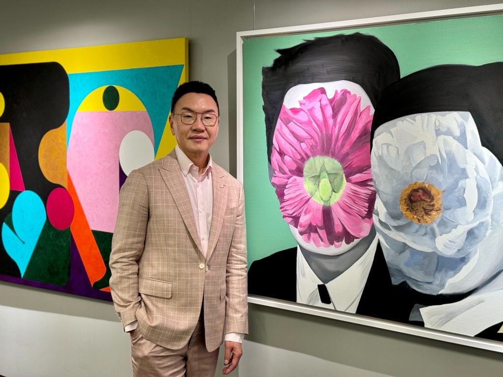 An East Asian man in a beige suit standing in front of colorful paintings