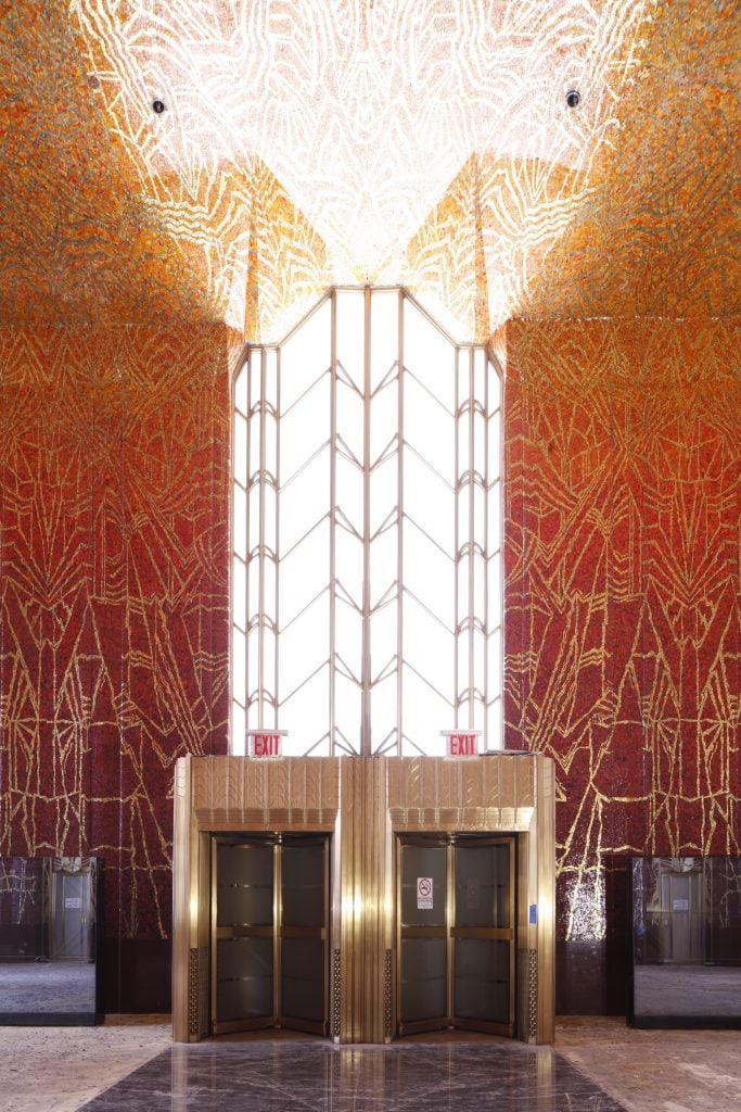 Light streams from a geometric shaped glass paneled window in a high-ceilinged room with ornate Art Deco mosaics by Hildreth Meière fading from blood red to bright orange on the ceiling, decorated with sparkling gold designs.