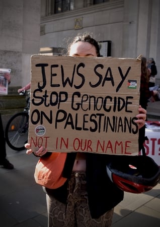 a person stands behind a cardboard sign on which is written "Jews say stop genocide on Palestine: not in our name"