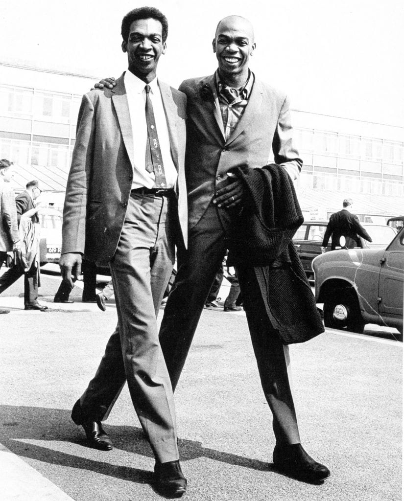 a black and white photograph of two men walking next two other, they are wearing suits