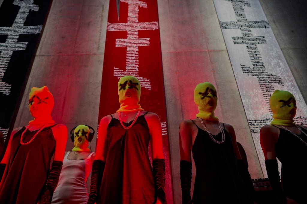 a group of women in black dresses and yellow balaclavas stand in front of vertical banners with symbols running down them