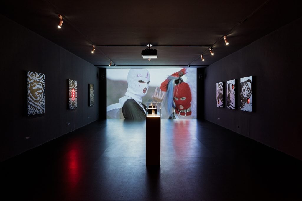 a large room with a video projected onto the far wall with two people in white and red balaclavas and a podium in the middle of the room holding a small glass vial
