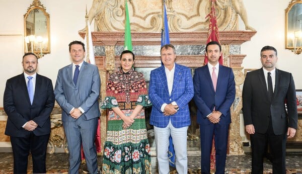 Men in suits and a woman in a long-sleeved dress with colorfully patterned stripes stand in a row in front of a fireplace with an ornate marble mantlepiece flanked by small gold-framed mirrors. 