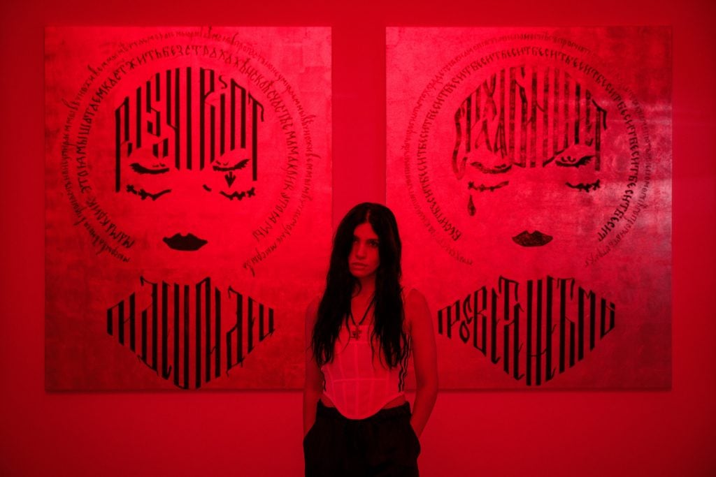 a red lit room in which a woman with brown hair stands in front of two images on the wall with vaguely human life faces (balaclavas) with text written over them in Russian 