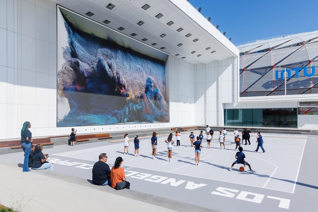 A large digital display showing an abstract, dynamic image resembling a wave or cloud, with people engaging in activities below.