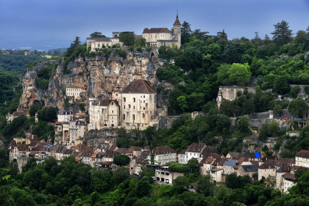 A present-day view from a distance of a Medieval town built in three levels on a cliff face, with red roofed light stone buildings and leafy green trees. 