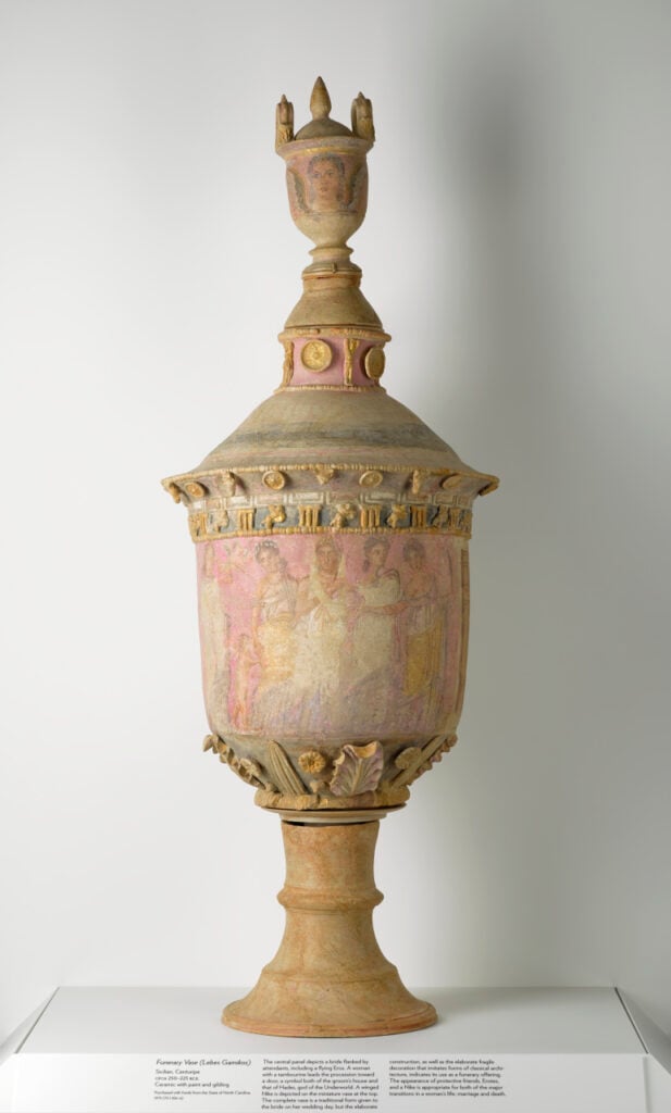 An ornate, ancient Sicilian vase used for funerary purposes, decorated with intricate carvings and figures, featuring a tall, elegant shape with a lid topped by a small statue.