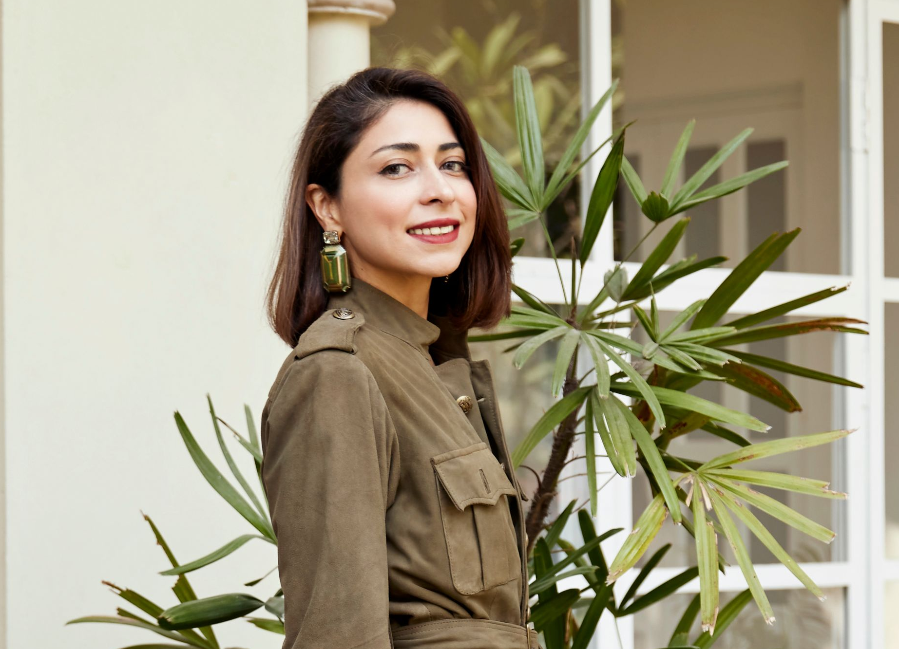 An Indian woman in dark brown shoulder length hair and a dark green jacket posing in front of a house