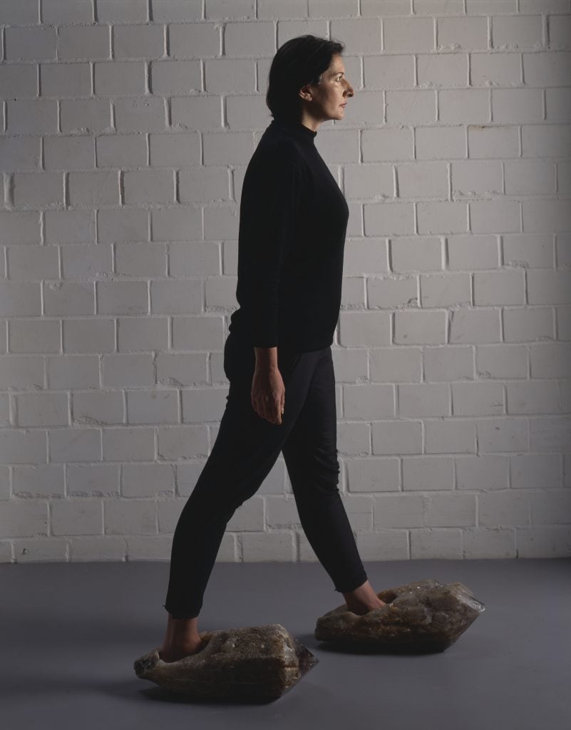 Marina Abramović, dressed in black, is seen with her feet inside large "shoes" made of quartz and amethyst crystal.