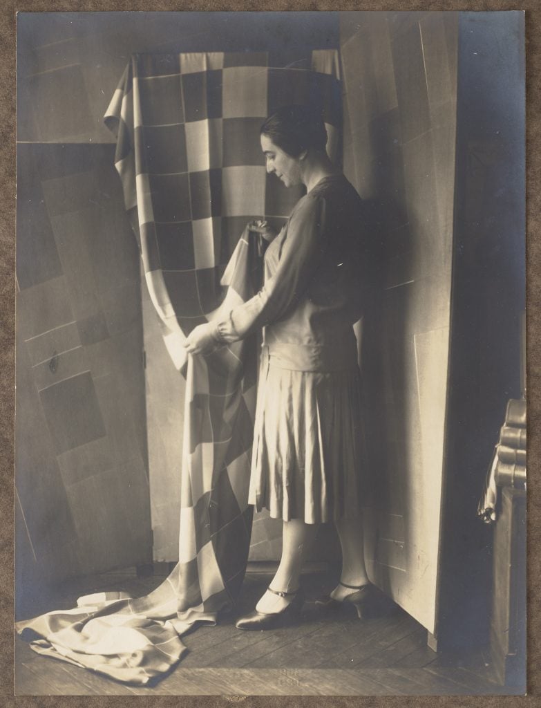 A black and white photograph of Sonia Delaunay in her studio at Boulevard Malesherbes, standing in front of a checked textile draped against a wall (ca. 1925). 