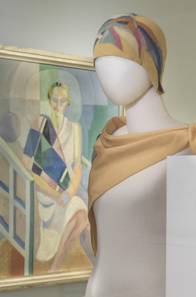A geometric looking painting by Robert Delaunay of a woman dressed in a white dress and colorful scarf behind a felted wool cloche and matching silk scarf by Sonia Delaunay on a mannequin bust on view at the Bard Graduate Center Gallery. 