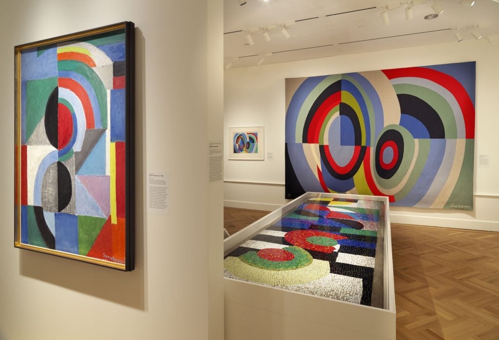 Installation view of "Sonia Delaunay: Living Art" is on view at the Bard Graduate Center Gallery featuring three works with a similar geometric design of concentric circles with geometric color blocking, one of a painting on the front wall, one in a mosaic displayed in a case, and the third of a tapestry hanging on the back wall next to a smaller study on paper of the same design. Photo ©Bruce M. White.