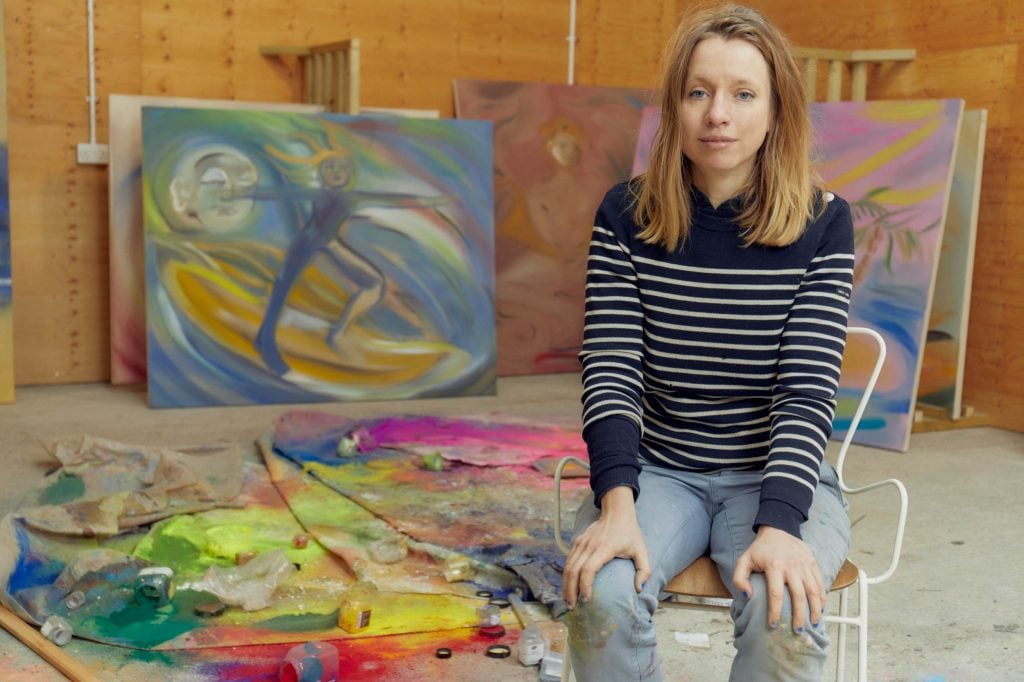 a woman in a navy and white striped jumper and jeans sits on a chair in front of large canvases and bits of paint, it is clearly an artist's studio