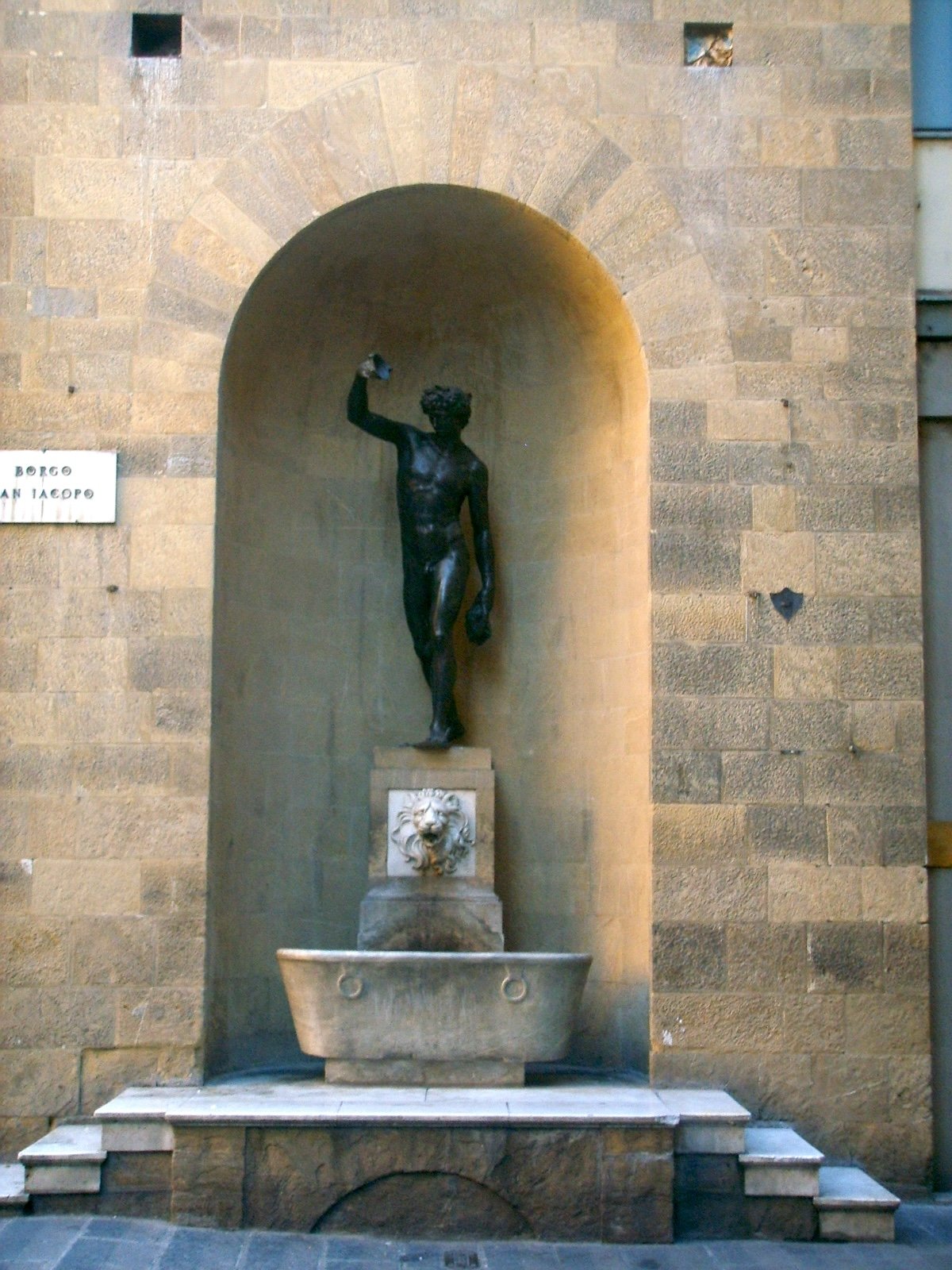 A bronze statue of Bacchus mounted on a marble fountain in a niche on a wall of the Torre dei Rossi-Cerchi, a rebuilt medieval tower tan stones in Florence.