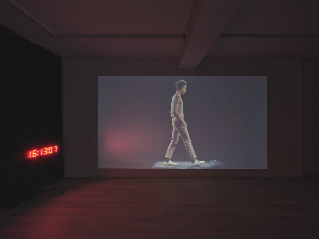 video installation showing a man walking on a tread mill beside a clock