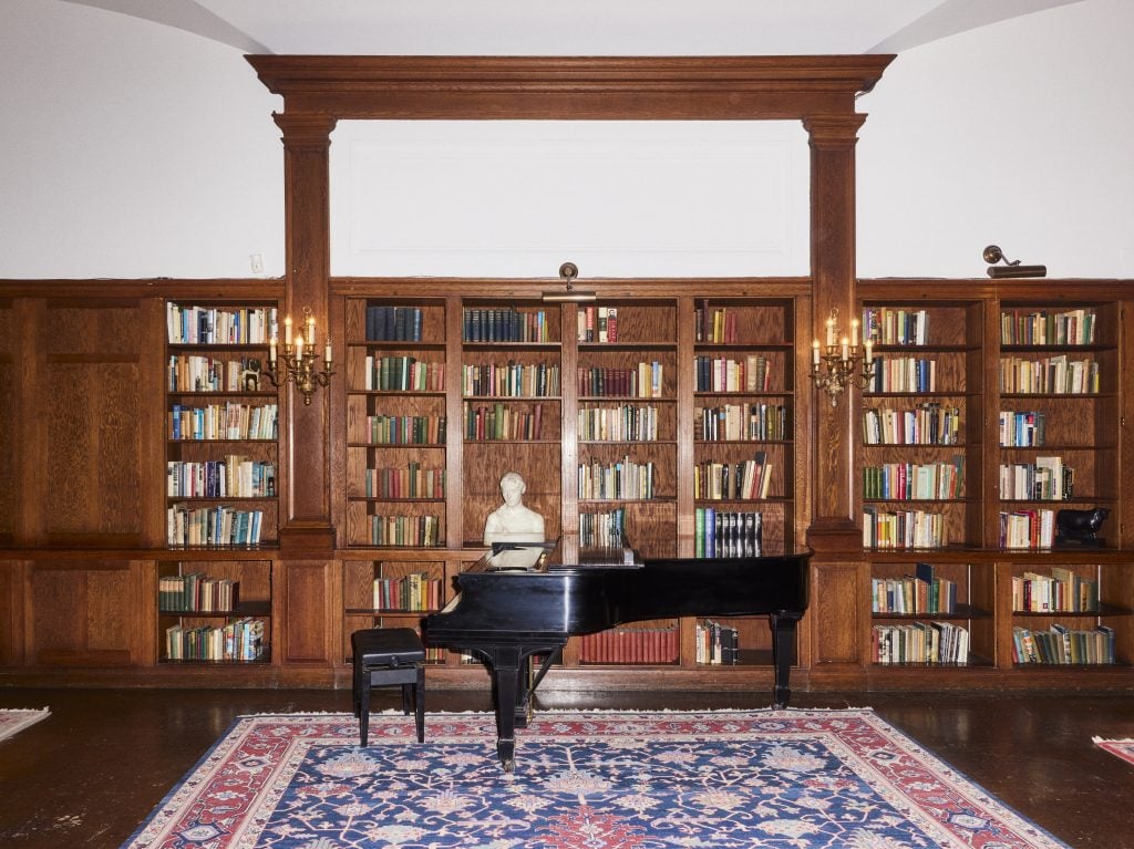 A library with built in book shelves and a black piano on a large red and blue woven carpet.
