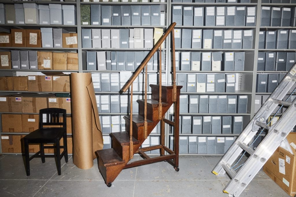 A room filled with gray file boxes behind a black chair, a roll of brown paper, a tall wooden step stool with handrails, and a silver ladder leaning against a cardboard box.