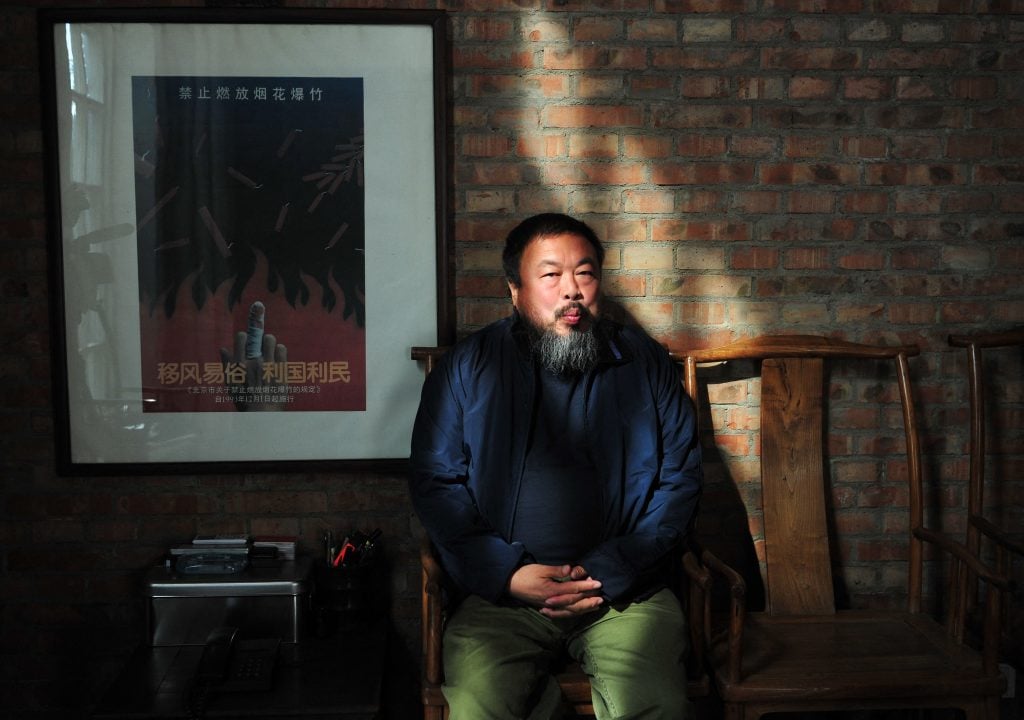 Artist Ai Weiwei sitting in a chair against a brick wall, a poster framed beside him