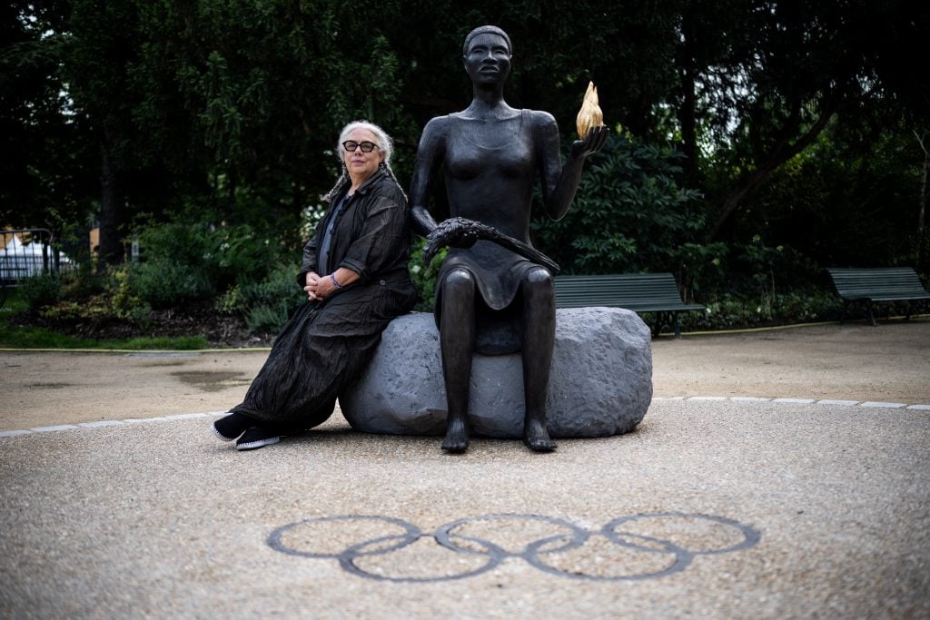 Artist Alison Saar, a fair Black woman with dark rimmed glasses and white hair in two long braids, sits in the park next to her sculpture The Salon, a new monument on the Champs-Élysées in Paris commissioned for the Olympic Games. The bronze artwork is a larger-than-life Black woman holding a golden flame. The Olympic rings are engraved in the ground in front of them.