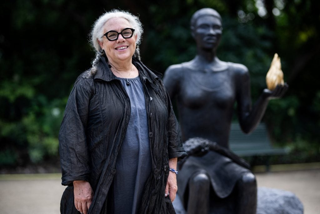 Artist Alison Saar, a fair Black woman with dark rimmed glasses and white hair in two long braids, stands in the park next to her sculpture The Salon, a new monument on the Champs-Élysées in Paris commissioned for the Olympic Games. The bronze artwork is a larger-than-life Black woman holding a golden flame.