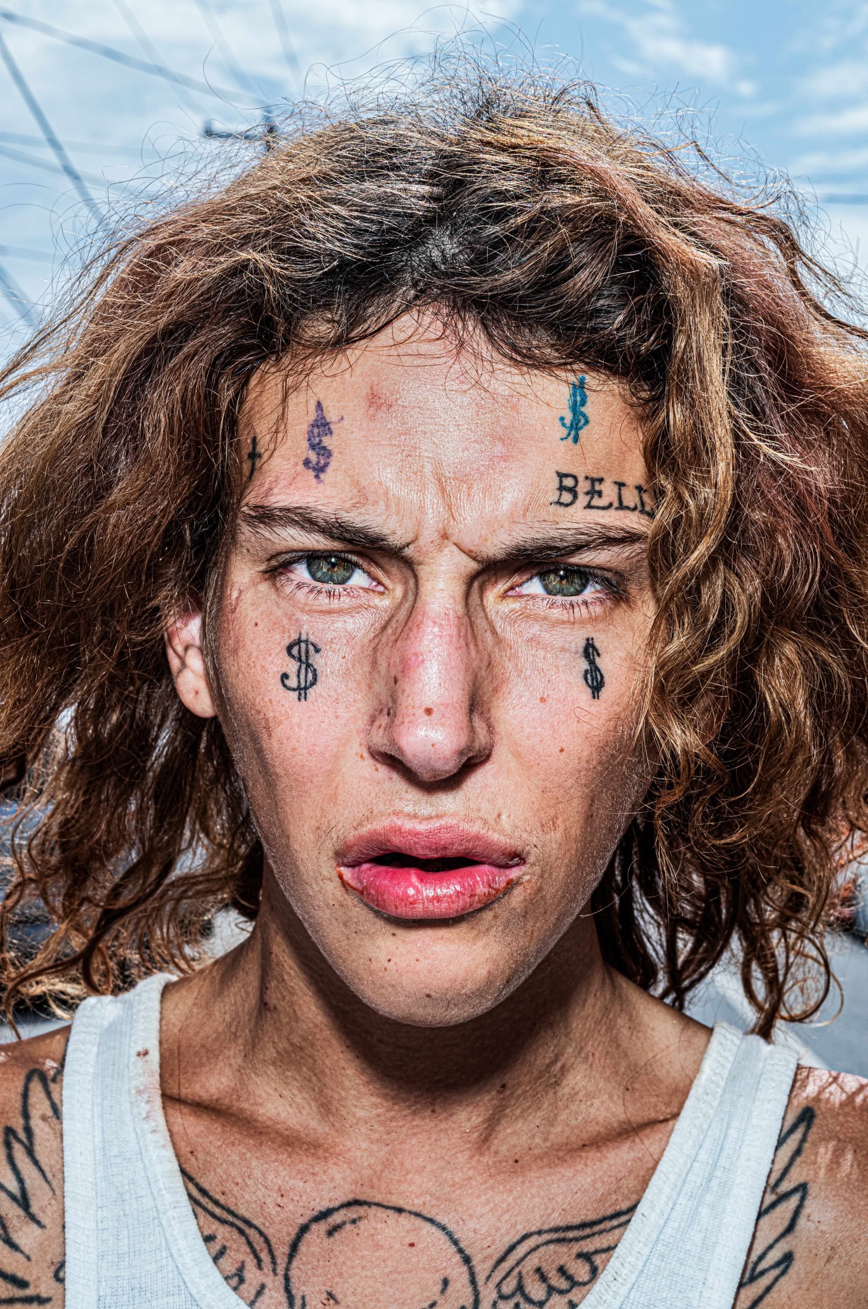 A close up color photo portrait of a white individual with wavy messy dark blonde hair and green eyes with four dollar sign tattoos on their face and the word "bell" over their left eyebrow, made by Bruce Gilden one of the street photographers being shown at Fotografiska.