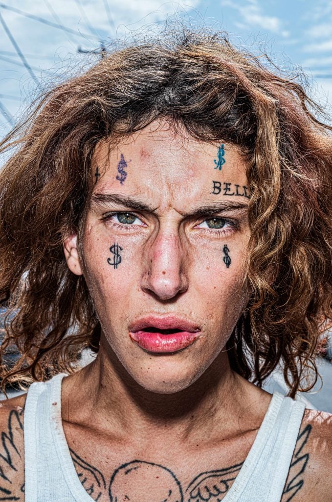 A close up color photo portrait of a white individual with wavy messy dark blonde hair and green eyes with four dollar sign tattoos on their face and the word 