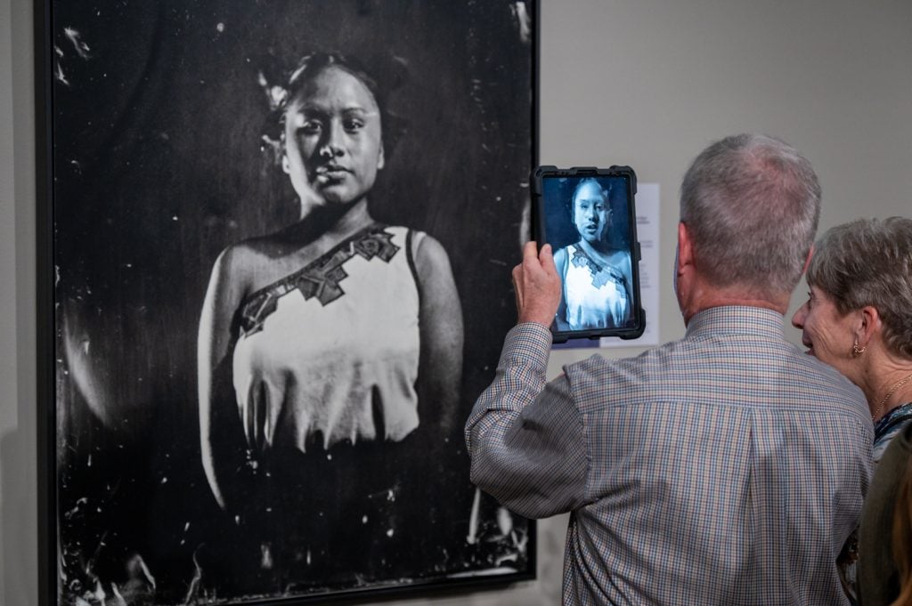 A black and white photograph of a young girl on the wall with two visitors to the exhibition staged by Art Bridges Foundation hold up an iPad to take a picture of it, and it can be seen on the device's screen.