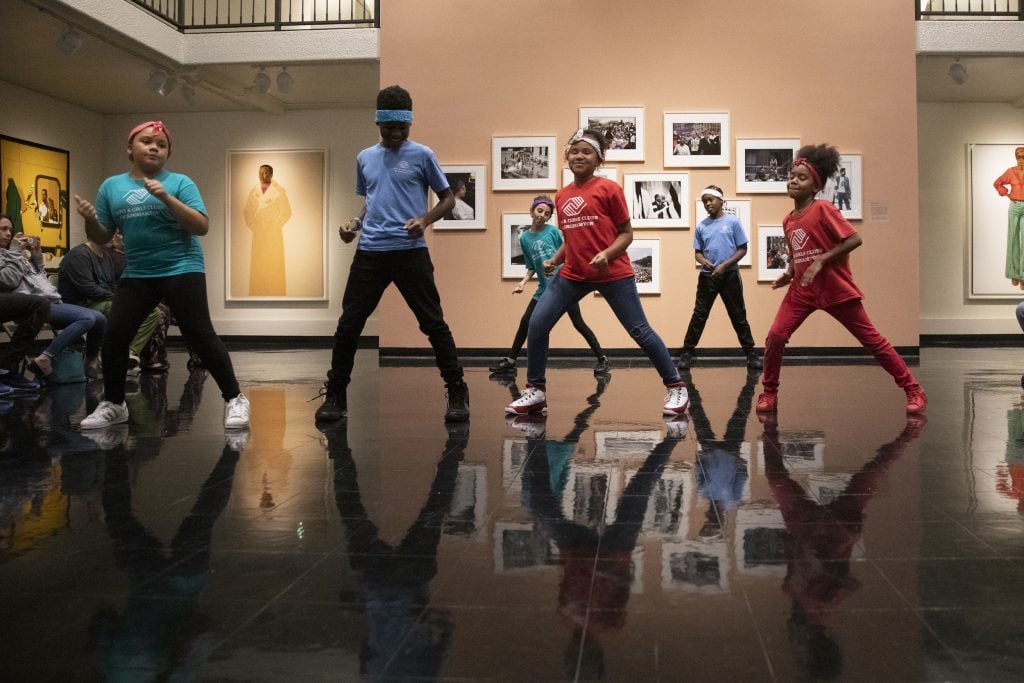 A group of tween children dance in the middle of a gallery space installed with salon style hung arworks on a reflective black floor within one of the art bridges foundation facilitated exhibitions.