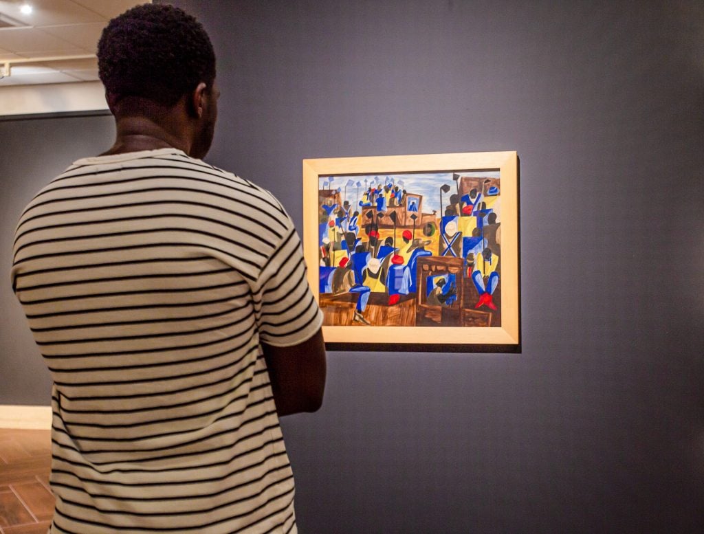 A man wearing a black and white striped tee looks at a painting installed on a dark purple wall.