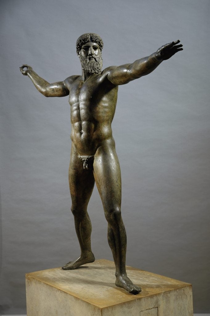 A patinad bronze sculpture of a a man standing widthwise with his left arm stretched forward and right arm pulled back with an opening in his hands that once would have held a trident or thunderbolt that was about to be thrown like a javelin, standing on a block of rough marble in front of a gray backdrop.
