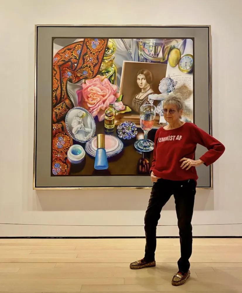 The elderly artist Audrey Flack dressed in tight black pants and a red sweatshirt reading "Feminist AF" stands in front of her Photorealist still life painting Leonardo's Lady in the Museum of Modern Art. The large painting shows the contents of a woman's nightstand, including a large pink rose, a bottle of blue nail polish, an open makeup compact, an open tub of facial cream, a Roman style statuette of a baby with grapes and a grape headdress, a wide black ribbon with orange embroidery, a small glass of water, a green pear, a small wine glass of water, a gold perfume bottle,a rhinestone brooch, and an art history text book open to a reproduction of Leonado da Vinci's painting La Belle Ferronnière.