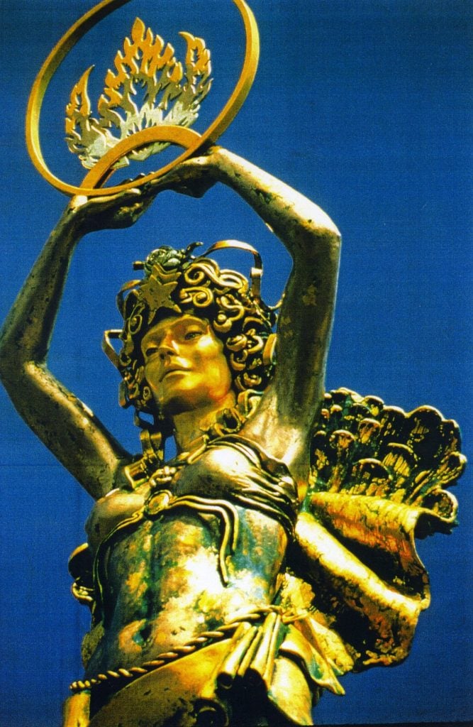 A gilded bronze statue of a female goddess with curly hair and a star diadem holds a ring with a flame symbol inside it aloft above her head is seen against a bright blue sky.  