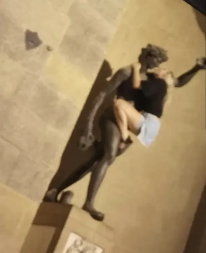 A blonde women in denim shorts and a black top embraces a nude bronze statue of Bacchus by Giambologna in a blurry photograph seemingly taken at night.