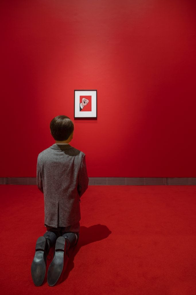 a model of a young boy in a suit kneels before a small painting on a red wall of a pointing finger