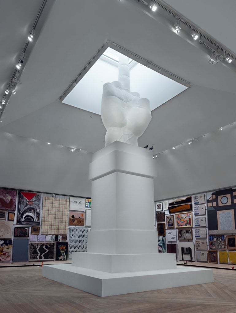 a glowing white hand with the middle finger up reaches up from a podium into a skylight inside a gallery space.
