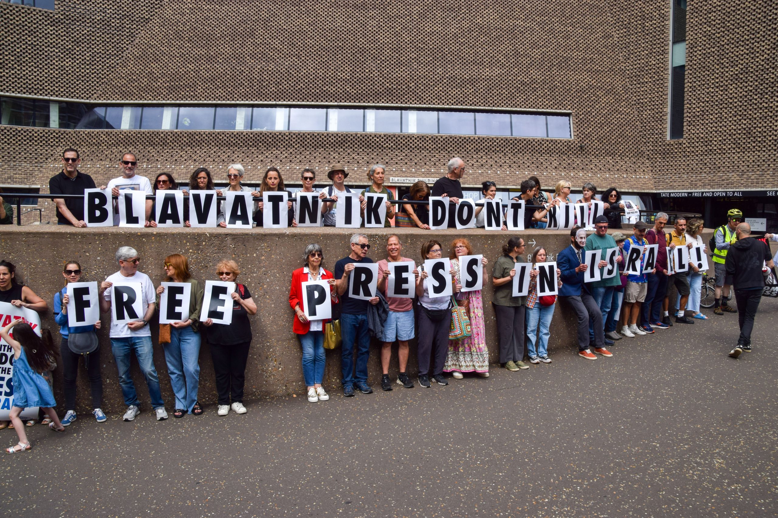 a line of protestors stand in a line holding up letters that spell out "FREE PRESS IN ISRAEL" in front of a brown building