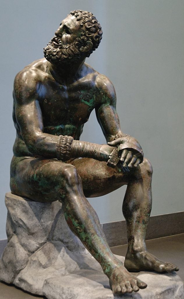 A patinad bronze sculpture of a man seated on a stone looking up and to his right wearing boxing hand wraps and with small gashes on his ears and curly hear and beard.