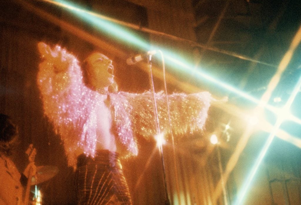 A man in a feathered shirt spreads his arms and sings into the mic amid colorful stage lights