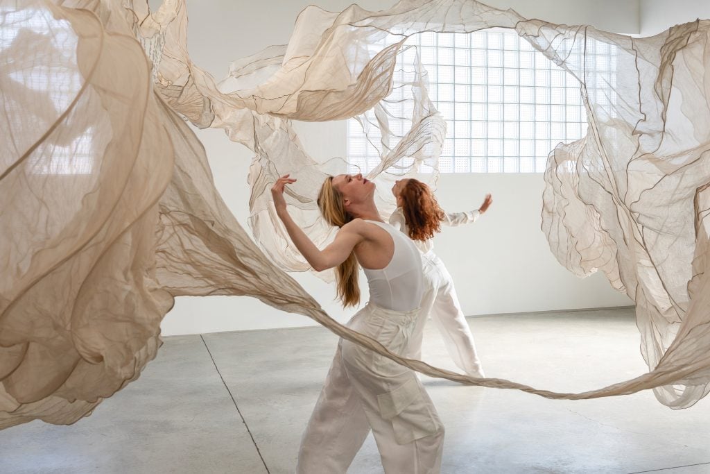 Two dancers dressed in white mid-performance within an off-white textile hanging installation artwork inside Brooklyn gallery Carvalho Park.