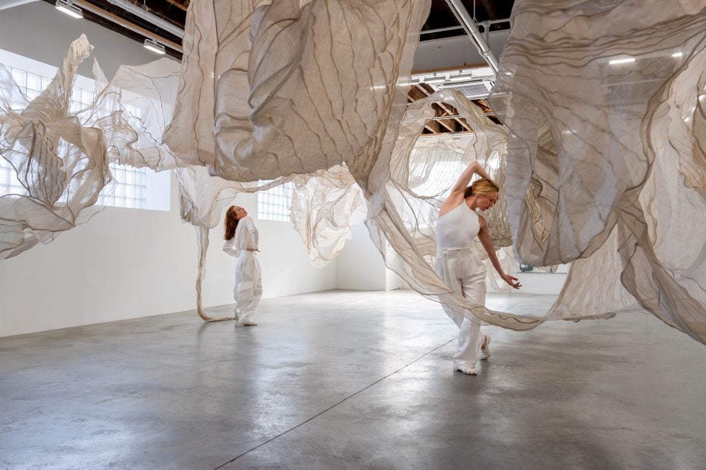 Sara Mearns and Jodi Melnick in the midst of a dance performance inside a textile installation of beige and cream hanging fabric at Carvahlo Park.